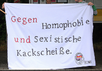 Foto eines Transparents. Darauf steht in roter und schwarzer Schrift: "Gegen Homophobie und sexistische Kackscheiße." Im unteren rechten Eck befindet sich das Logo der Antihomophoben Aktion Bonn.